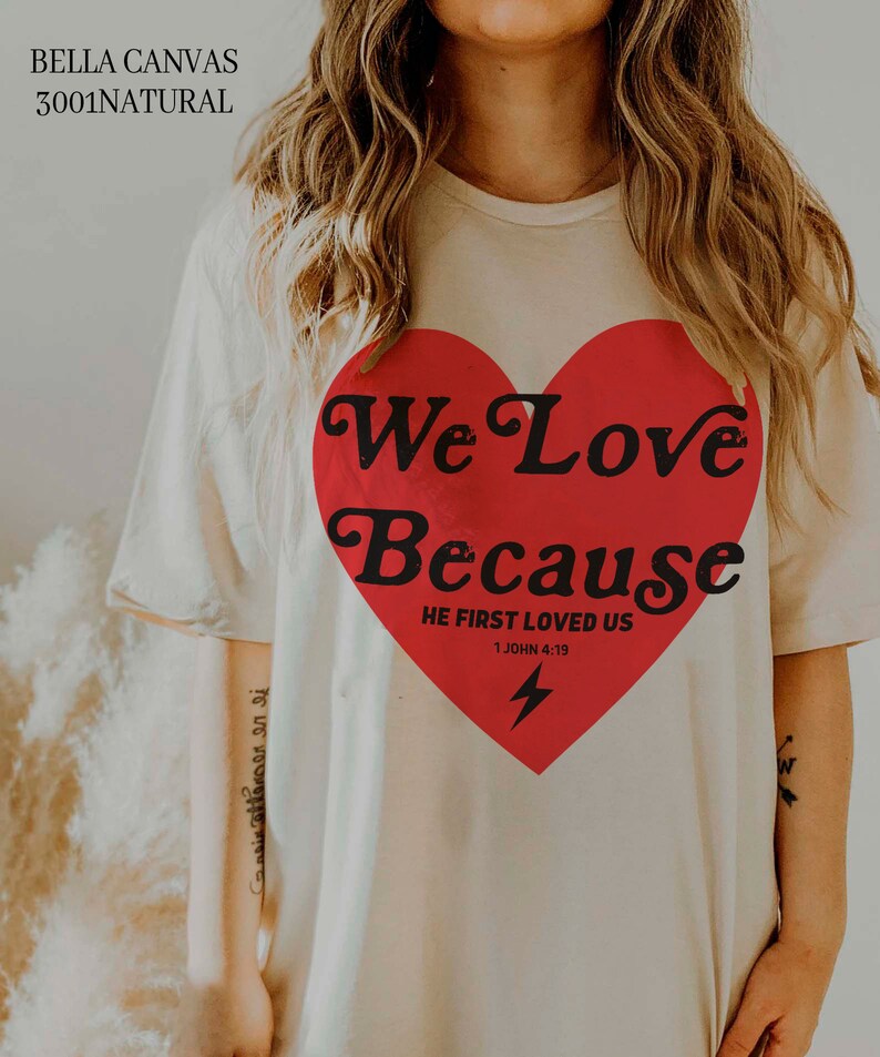May include: Cream-colored t-shirt with a large red heart graphic. The heart contains the words "We Love Because" and "He First Loved Us 1 John 4:19" in black text, along with a lightning bolt. The shirt is made of Bella Canvas 3001 Natural material.
