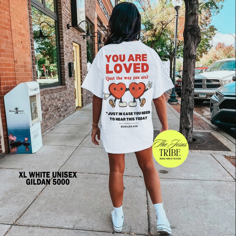 K&ouml;nnte beinhalten: Wei&szlig;es Unisex-T-Shirt mit einem Grafikdruck von zwei roten Herzen mit Gesichtern und dem Text "You are loved (just the way you are)". Der Text "Just in case you need to hear this today Romans 8:38" befindet sich unter den Herzen. Der Text "The Jesus Tribe" befindet sich am unteren Rand des T-Shirts.