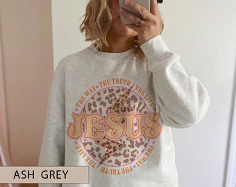 May include: Pale grey sweatshirt with a circular design. The design includes the word "JESUS" in a retro font, a lightning bolt, and the phrases "THE WAY THE TRUTH THE LIFE". The design has a leopard print background.