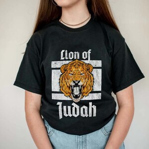 May include: Black t-shirt featuring a distressed graphic of a roaring lion's head in shades of orange and brown, set within a white rectangular frame. The words "Lion of Judah" are printed in a vintage font below the lion image.