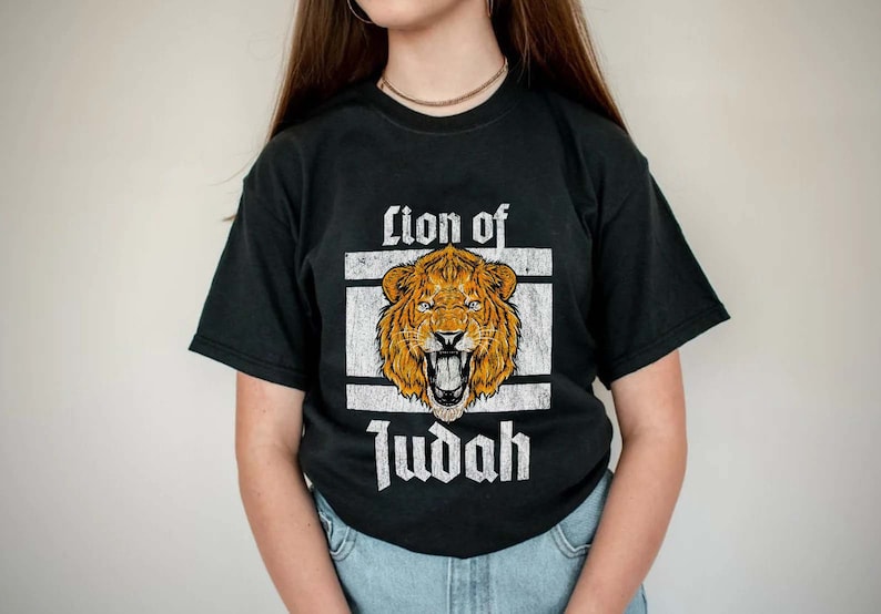 May include: Black t-shirt featuring a distressed graphic of a roaring lion's head in shades of orange and brown, set within a white rectangular frame. The words "Lion of Judah" are printed in a vintage font below the lion image.