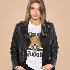 May include: A white t-shirt with a graphic of a lion's head and the words "Lion of Judah" in black. The shirt is worn under a black leather jacket and black pants. The background is a solid tan color.