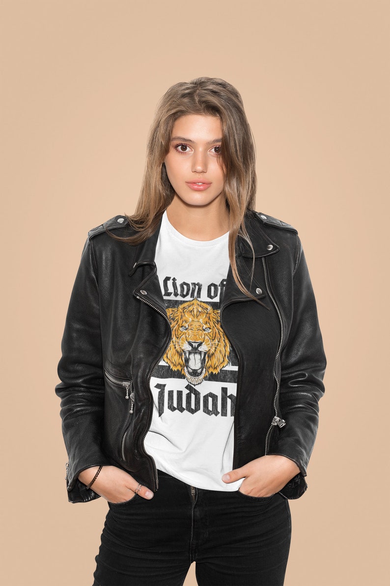May include: A white t-shirt with a graphic of a lion's head and the words "Lion of Judah" in black. The shirt is worn under a black leather jacket and black pants. The background is a solid tan color.