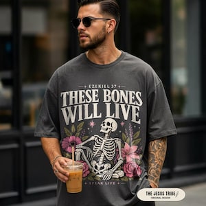 May include: Dark grey t-shirt with a skeleton graphic and the text "THESE BONES WILL LIVE". The design includes pink flowers and the text "+ EZEKIEL 37 +" and "+ SPEAK LIFE +".