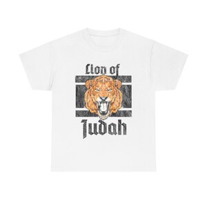 May include: White t-shirt featuring a distressed graphic of a roaring lion with the text "Lion of Judah" in a bold, vintage font. The lion is depicted in shades of orange and brown, set against a black and white rectangular frame.