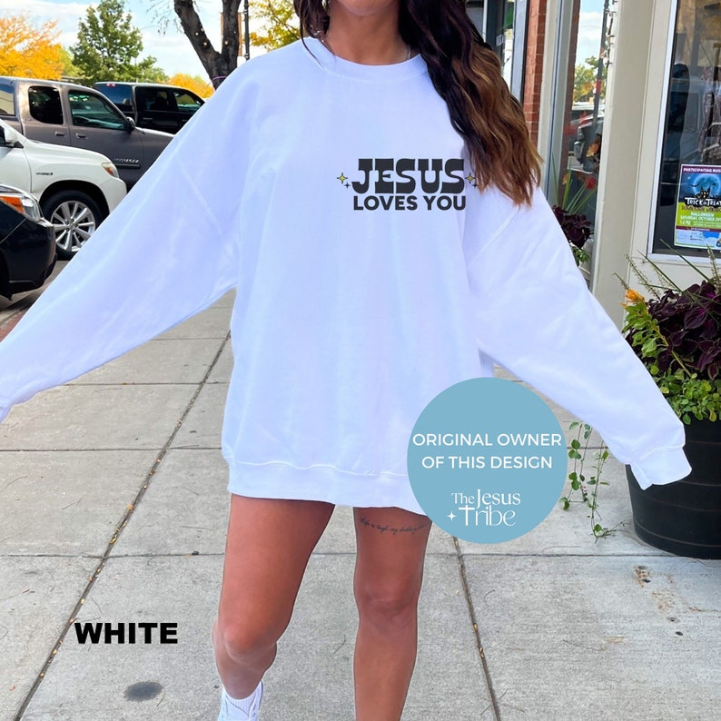 K&ouml;nnte beinhalten: Wei&szlig;es Sweatshirt mit dem schwarzen Text "JESUS LOVES YOU" und Sternen. Das Design stammt von "The Jesus + Tribe". Das Wort "WHITE" befindet sich am unteren Bildrand.