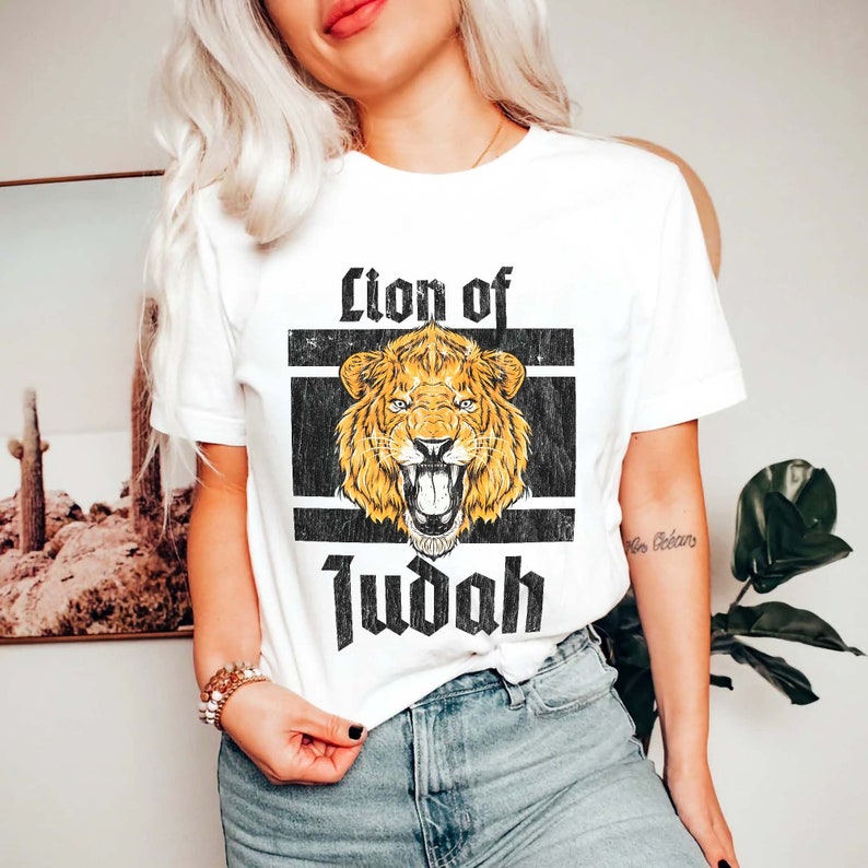 May include: White t-shirt featuring a distressed graphic of a roaring lion with the text "Lion of Judah" in a bold, gothic font. The lion is golden yellow, set against a black and white rectangular background.