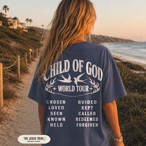 Op de afbeelding: Een blauw T-shirt met korte mouwen en witte tekst op de achterkant met de tekst CHILD OF GOD WORLD TOUR, twee vogelillustraties en een lijst met woorden: CHOSEN, GUIDED, LOVED, KEPT, SEEN, CALLED, KNOWN, REDEEMED, HELD en FORGIVEN.