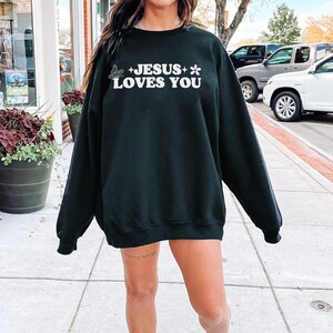 May include: Black sweatshirt with the text "JESUS LOVES YOU" in white, along with a butterfly and flower graphic. The sweatshirt is a crew neck style and appears to be made of a soft material.