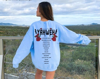 May include: A light blue sweatshirt with the word "YAHWEH" printed in black with lightning bolts on either side. The text is surrounded by red roses and a list of titles and descriptions in black text, including "Alpha and Omega", "King of Kings", "Lord of Lords", "The Great I Am", "Firstborn", "Father", "Healer", "Prince of Peace", "Redeemer", "Lion and the Lamb", "Savior", "Shepherd", and "Creator".
