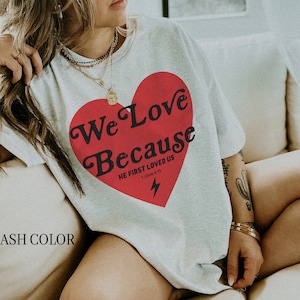 May include: Ash-colored t-shirt featuring a large red heart with the words "We Love Because" and "He First Loved Us" printed in black. A lightning bolt graphic is below the text. The shirt is a casual wear item.