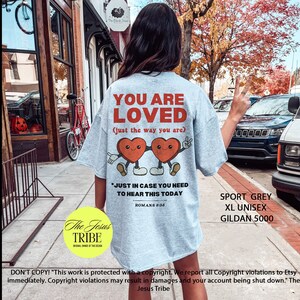 K&ouml;nnte beinhalten: Ein hellgraues T-Shirt mit dem Text "You Are Loved (just the way you are)" und zwei Comic-Herzen, die sich an den H&auml;nden halten. Der Text "Just in case you need to hear this today" befindet sich unter den Herzen, mit dem Schriftzitat "R&ouml;mer 8:38" darunter. Das T-Shirt hat auch den Text "Sport Grey XL Unisex Gildan 5000" in der unteren rechten Ecke.