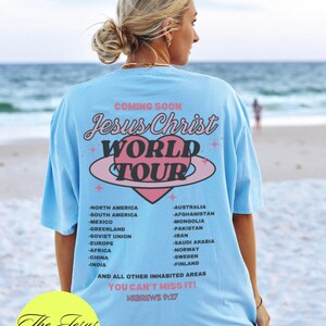 Puede incluir: Una camiseta azul claro con un gr&aacute;fico de coraz&oacute;n rosa y el texto "Jesus Christ World Tour" impreso en la espalda. El texto "You Can't Miss It! Hebrews 9:27" tambi&eacute;n est&aacute; impreso en la camiseta. La camiseta la lleva puesta una persona de pie en una playa.