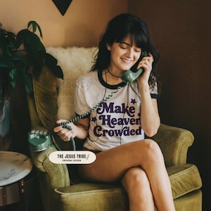 Op de afbeelding: Een wit ringer-t-shirt met donkerblauwe boorden aan de mouwen en halslijn. Het shirt heeft de paarse tekst "Make Heaven Crowded" met kleine stergraphics. Een groene vintage draaitelefoonhoorn wordt met een snoer tegen het oor gehouden.