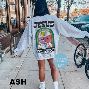 K&ouml;nnte beinhalten: Hellgraues Sweatshirt mit dem Text "JESUS LOVES YOU (AND I'M TRYING)" &uuml;ber einer Cartoon-Grafik eines Herzens und eines Pilzes in einem Regenbogen. Das Design enth&auml;lt den Text "ORIGINAL OWNER OF THIS DESIGN The Jesus + Tribe".