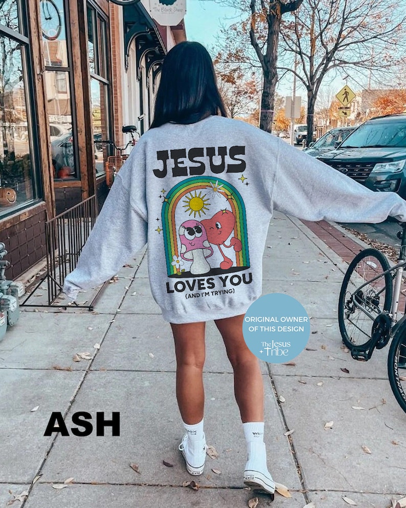 K&ouml;nnte beinhalten: Hellgraues Sweatshirt mit dem Text "JESUS LOVES YOU (AND I'M TRYING)" &uuml;ber einer Cartoon-Grafik eines Herzens und eines Pilzes in einem Regenbogen. Das Design enth&auml;lt den Text "ORIGINAL OWNER OF THIS DESIGN The Jesus + Tribe".