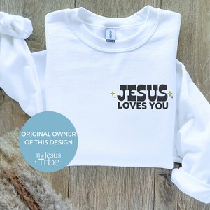 K&ouml;nnte beinhalten: Wei&szlig;es Sweatshirt mit dem Text "JESUS LOVES YOU" in schwarzen Blockbuchstaben. Das Design enth&auml;lt Sternakzente. Das Sweatshirt ist auf einer hellen Holzoberfl&auml;che gefaltet. Der Text "ORIGINAL OWNER OF THIS DESIGN" ist ebenfalls sichtbar.