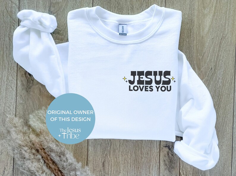 K&ouml;nnte beinhalten: Wei&szlig;es Sweatshirt mit dem Text "JESUS LOVES YOU" in schwarzen Blockbuchstaben. Das Design enth&auml;lt Sternakzente. Das Sweatshirt ist auf einer hellen Holzoberfl&auml;che gefaltet. Der Text "ORIGINAL OWNER OF THIS DESIGN" ist ebenfalls sichtbar.