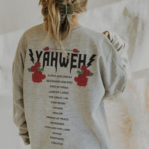May include: Grey sweatshirt with the word "YAHWEH" printed in black with lightning bolts on either side. The word is surrounded by red roses. Below the word are the words "ALPHA AND OMEGA", "BEGINNING AND END", "KING OF KINGS", "LORD OF LORDS", "THE GREAT I AM", "FIRSTBORN", "FATHER", "HEALER", "PRINCE OF PEACE", "REDEEMER", "LION AND THE LAMB", "SAVIOR", "SHEPHERD", "CREATOR".