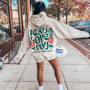 K&ouml;nnte beinhalten: Ein cremefarbener Hoodie mit einer gr&uuml;nen und pinkfarbenen Grafik, auf der "hey! JESUS LOVES YOU" und "OKAY, HAVE A GREAT DAY, BY THE JESUS TRIBE CHRISTIAN APPAREL" steht.