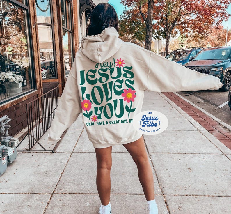 K&ouml;nnte beinhalten: Ein cremefarbener Hoodie mit einer gr&uuml;nen und pinkfarbenen Grafik, auf der "hey! JESUS LOVES YOU" und "OKAY, HAVE A GREAT DAY, BY THE JESUS TRIBE CHRISTIAN APPAREL" steht.
