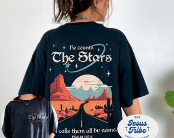 May include: Black t-shirt with a graphic of a desert landscape with a road leading to a moon and stars. The text "He counts The Stars" is above the graphic and "calls them all by name. PSALM 147:4" is below. The text "THE Jesus +Tribe+ CHRISTIAN APPAREL" is in a white oval on the bottom right corner.