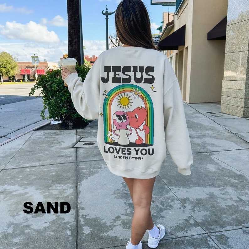 K&ouml;nnte beinhalten: Sandfarbenes Sweatshirt mit dem Text "JESUS LOVES YOU (AND I'M TRYING)". Auf der R&uuml;ckseite des Sweatshirts befindet sich eine farbenfrohe Grafik eines Pilzes und eines Herzens in einem Regenbogenbogen. Das Sweatshirt hat eine lockere Passform.