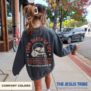 K&ouml;nnte beinhalten: Dunkelgraues Sweatshirt mit dem Text "FROM DEATH TO LIFE" &uuml;ber einer Totenkopf-Grafik. Auf der R&uuml;ckseite des Sweatshirts befindet sich auch ein Bibelvers. Die linke untere Ecke des Bildes tr&auml;gt den Text "COMFORT COLORS".
