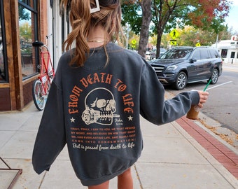 May include: Dark gray sweatshirt with the text "FROM DEATH TO LIFE" above a skull graphic. The back of the sweatshirt also features a Bible verse. The bottom left corner of the image has the text "COMFORT COLORS".