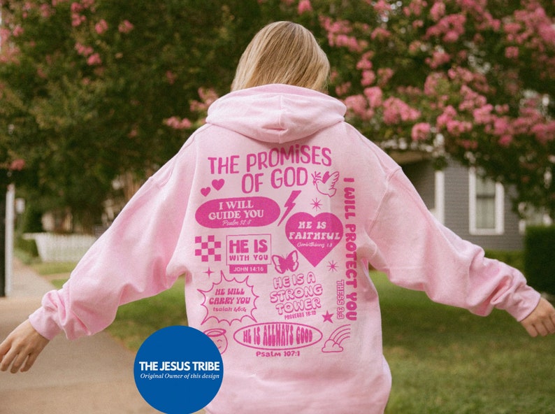 May include: A light pink hoodie with the text "THE PROMISES OF GOD" and various phrases and graphics in pink. The phrases include "I WILL GUIDE YOU", "HE IS WITH YOU", and "HE IS FAITHFUL". The hoodie also features the text "THE JESUS TRIBE".