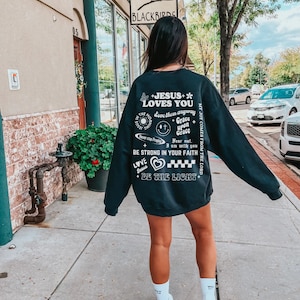 May include: A black sweatshirt with white text and graphics. The text includes "JESUS LOVES YOU", "BE STRONG IN YOUR FAITH", and other inspirational phrases. A butterfly, heart, and smiley face are also featured.