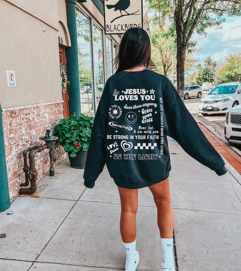 May include: A black sweatshirt with white text and graphics. The text includes "JESUS LOVES YOU", "BE STRONG IN YOUR FAITH", and other inspirational phrases. A butterfly, heart, and smiley face are also featured.