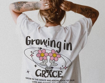 May include: White t-shirt with the text "Growing in GRACE" and a graphic of two pink flowers with smiley faces. The shirt also features the text "GROW IN THE GRACE AND KNOWLEDGE OF OUR LORD AND SAVIOR JESUS CHRIST" and "GROWTH IS A PROCESS".