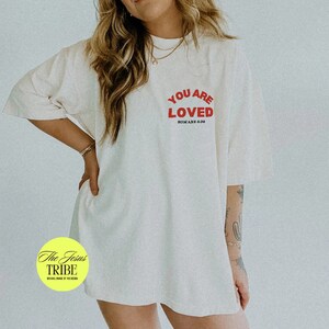 K&ouml;nnte beinhalten: Ein wei&szlig;es Oversize-T-Shirt mit rotem Text, der "You Are Loved" und "R&ouml;mer 8:38" in kleinerer Schrift lautet. Das T-Shirt wird von einer Person mit langen blonden Haaren getragen.