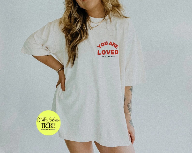 K&ouml;nnte beinhalten: Ein wei&szlig;es Oversize-T-Shirt mit rotem Text, der "You Are Loved" und "R&ouml;mer 8:38" in kleinerer Schrift lautet. Das T-Shirt wird von einer Person mit langen blonden Haaren getragen.