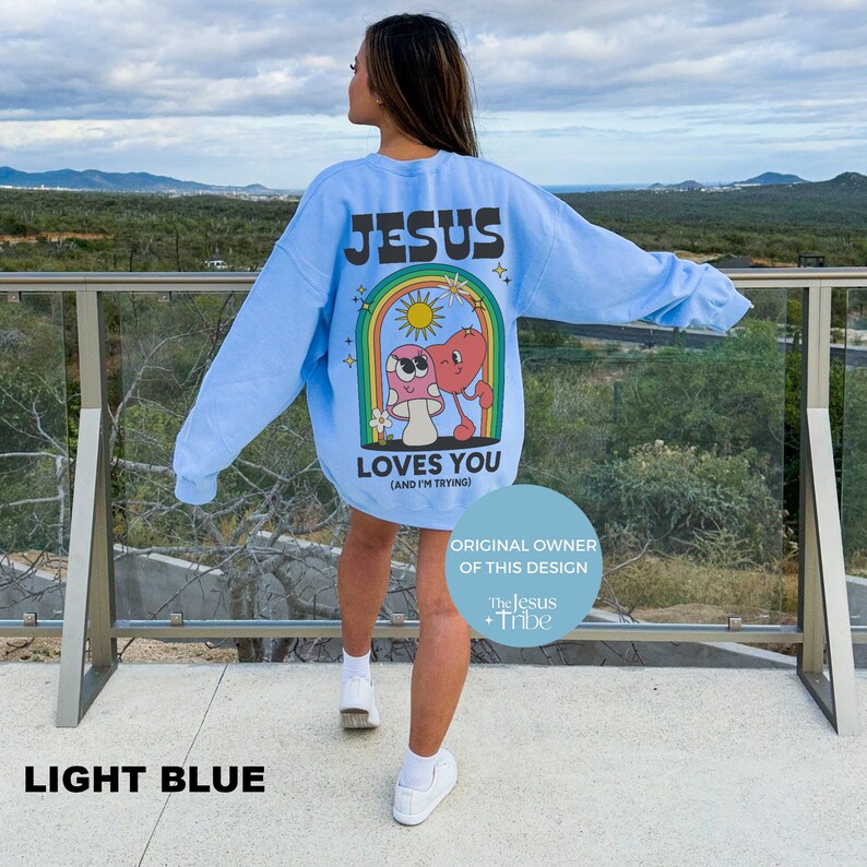 K&ouml;nnte beinhalten: Hellblaues Sweatshirt mit dem Text "JESUS LOVES YOU (AND I'M TRYING)" und einer Grafik aus Pilz und Herz unter einem Regenbogen. Das Sweatshirt ist hellblau.