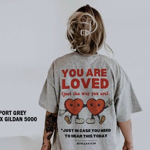 K&ouml;nnte beinhalten: Ein graues T-Shirt mit dem Text "YOU ARE LOVED (just the way you are)" in Rot. Das Shirt zeigt ein Cartoon-Bild von zwei roten Herzen mit Gesichtern und Armen, die sich die H&auml;nde halten. Der Text "*JUST IN CASE YOU NEED TO HEAR THIS TODAY ROMANS 8.38" ist unter dem Bild gedruckt.