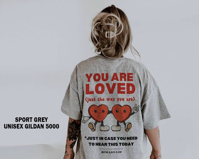 K&ouml;nnte beinhalten: Ein graues T-Shirt mit dem Text "YOU ARE LOVED (just the way you are)" in Rot. Das Shirt zeigt ein Cartoon-Bild von zwei roten Herzen mit Gesichtern und Armen, die sich die H&auml;nde halten. Der Text "*JUST IN CASE YOU NEED TO HEAR THIS TODAY ROMANS 8.38" ist unter dem Bild gedruckt.