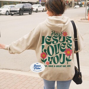 K&ouml;nnte beinhalten: Ein beiger Hoodie mit einer gr&uuml;nen und pinken Grafik, auf der "Hey! Jesus Loves You" steht, und einem kleinen Blumendesign. Der Hoodie hat auch einen wei&szlig;en ovalen Aufkleber mit dem Text "The Jesus Tribe Christian Apparel".