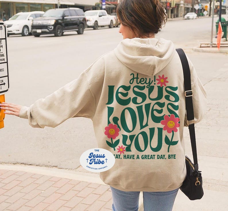 K&ouml;nnte beinhalten: Ein beiger Hoodie mit einer gr&uuml;nen und pinken Grafik, auf der "Hey! Jesus Loves You" steht, und einem kleinen Blumendesign. Der Hoodie hat auch einen wei&szlig;en ovalen Aufkleber mit dem Text "The Jesus Tribe Christian Apparel".