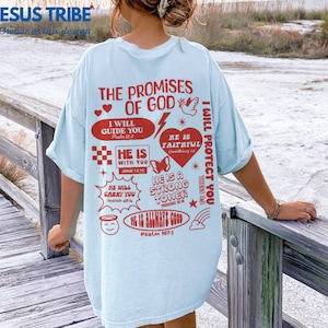 May include: Light blue t-shirt with a graphic print of religious quotes and symbols in red. The text includes "The Promises of God", "I Will Guide You", "He Is With You", "He Is Faithful", "He Is A Strong Tower", "I Will Protect You", and "He Is Always Good".