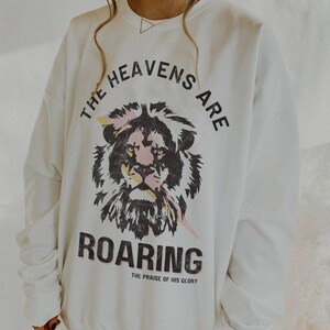 May include: White sweatshirt featuring a lion graphic with pink and yellow accents, and the text "THE HEAVENS ARE ROARING" in black. The sweatshirt is labelled "XL WHITE UNISEX GILDAN 1800".