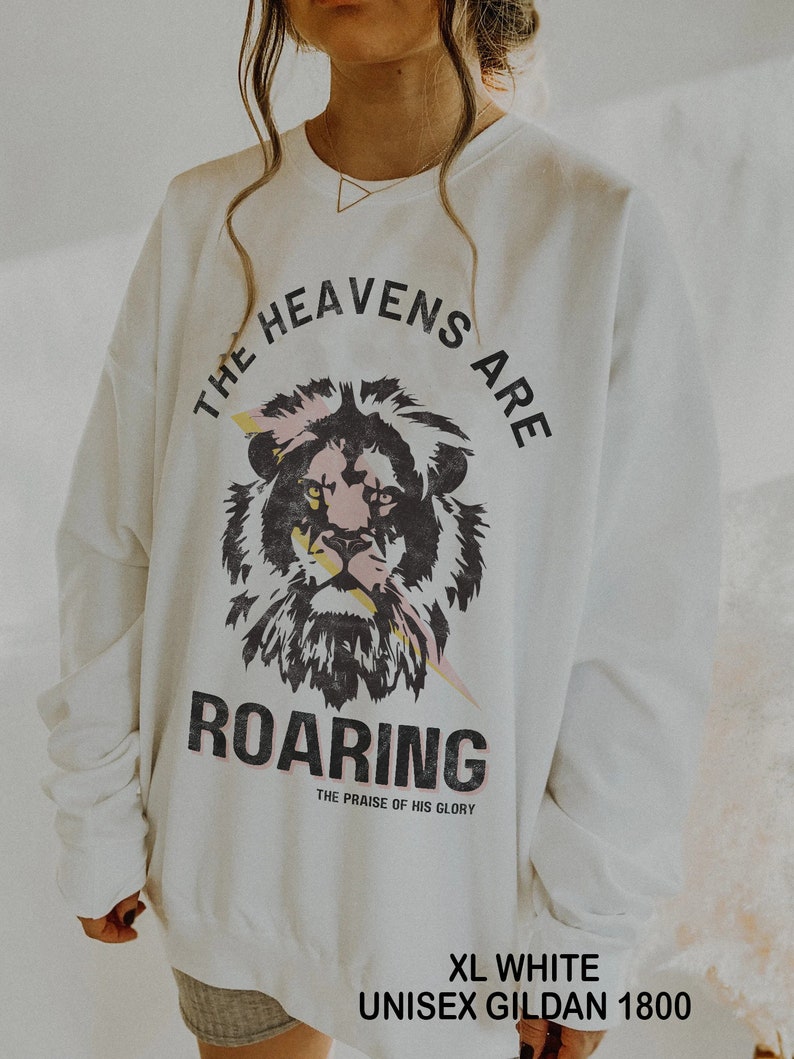 May include: White sweatshirt featuring a lion graphic with pink and yellow accents, and the text "THE HEAVENS ARE ROARING" in black. The sweatshirt is labelled "XL WHITE UNISEX GILDAN 1800".