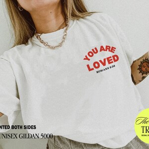 K&ouml;nnte beinhalten: Wei&szlig;es Unisex-T-Shirt mit dem Text "YOU ARE LOVED" in roten Buchstaben und dem Text "ROMANS 8:38" in schwarzen Buchstaben. Das Shirt ist beidseitig bedruckt.