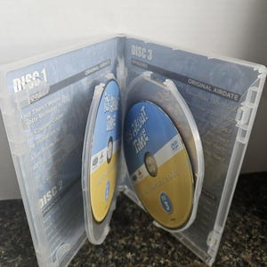 May include: An open clear plastic DVD case containing multiple discs for the television series It's About Time. The discs are blue and yellow with the show title printed on them. The interior panels list episode titles and original air dates.