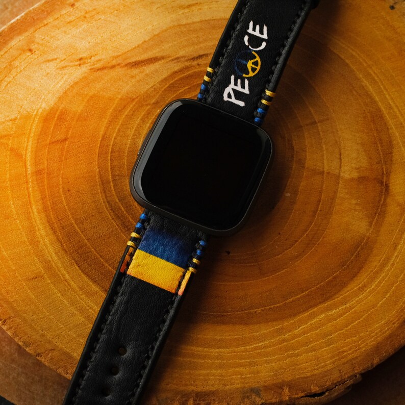 Puede incluir: Un reloj inteligente negro con una banda estampada en azul, amarillo y negro. La banda tiene la palabra "PEACE" bordada.
