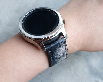 Peut inclure: Une montre connectée argentée avec un écran noir rond et un bracelet en cuir noir texturé et vieilli. La montre présente une lunette métallique avec des graduations et deux boutons latéraux, fixée par des coutures visibles sur les bords.