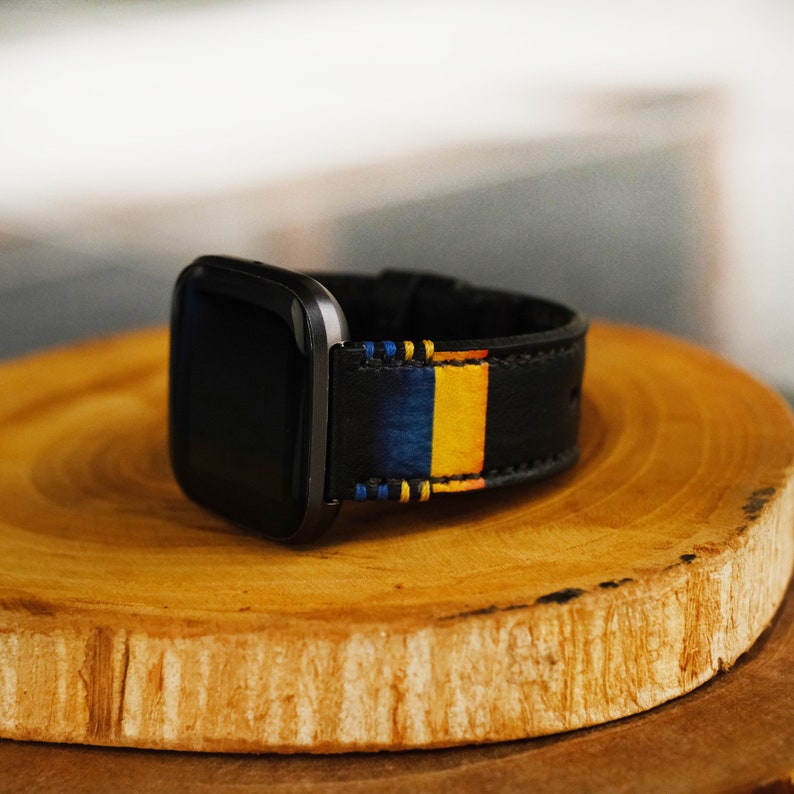 Puede incluir: Una correa de reloj de cuero negro con rayas azules, amarillas y blancas. La correa est&aacute; sobre una superficie de madera.