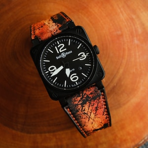 May include: A black Bell & Ross watch with a square face and orange and black patterned leather strap.