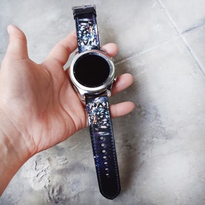 Op de afbeelding: Een smartwatch met een zilverkleurige, ronde metalen behuizing en een zwart digitaal display. De donkere leren horlogeband is voorzien van een gedetailleerde, kleurrijke grafische print van een wolvenkop op beide delen van de band.